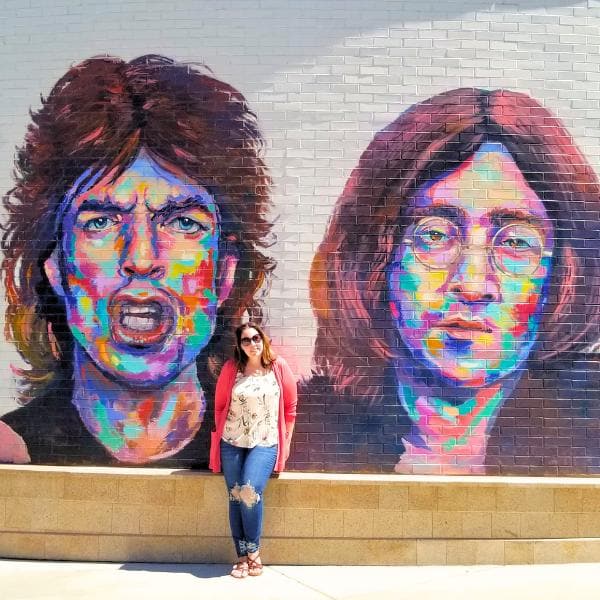 Girl standing in front of a brightly colored street art mural of musicians Mick Jagger and John Lennon