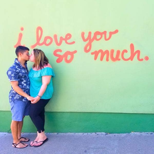 A couple holds hands and kisses in front of a green mural with "I Love You So Much" in orange cursive